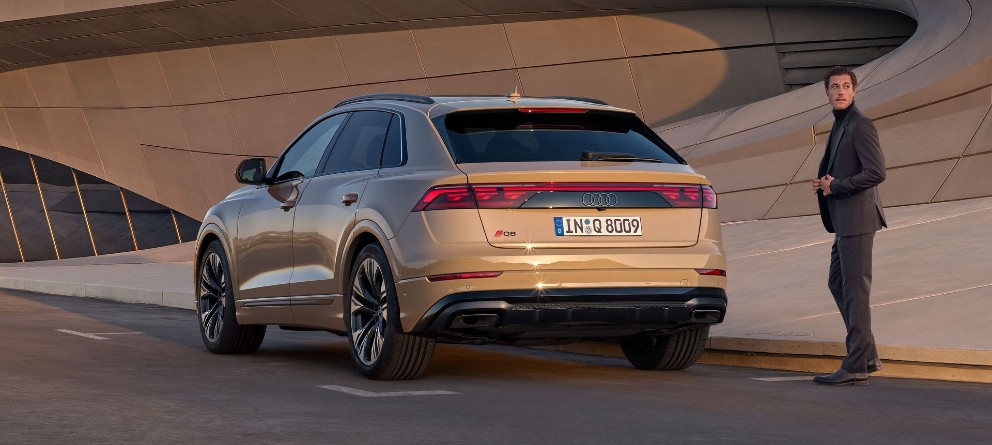 Audi Q8 marrón aparcado con un hombre elegante de mediana edad vestido de traje informal dirigiéndose hacia él. | eligetucoche.es